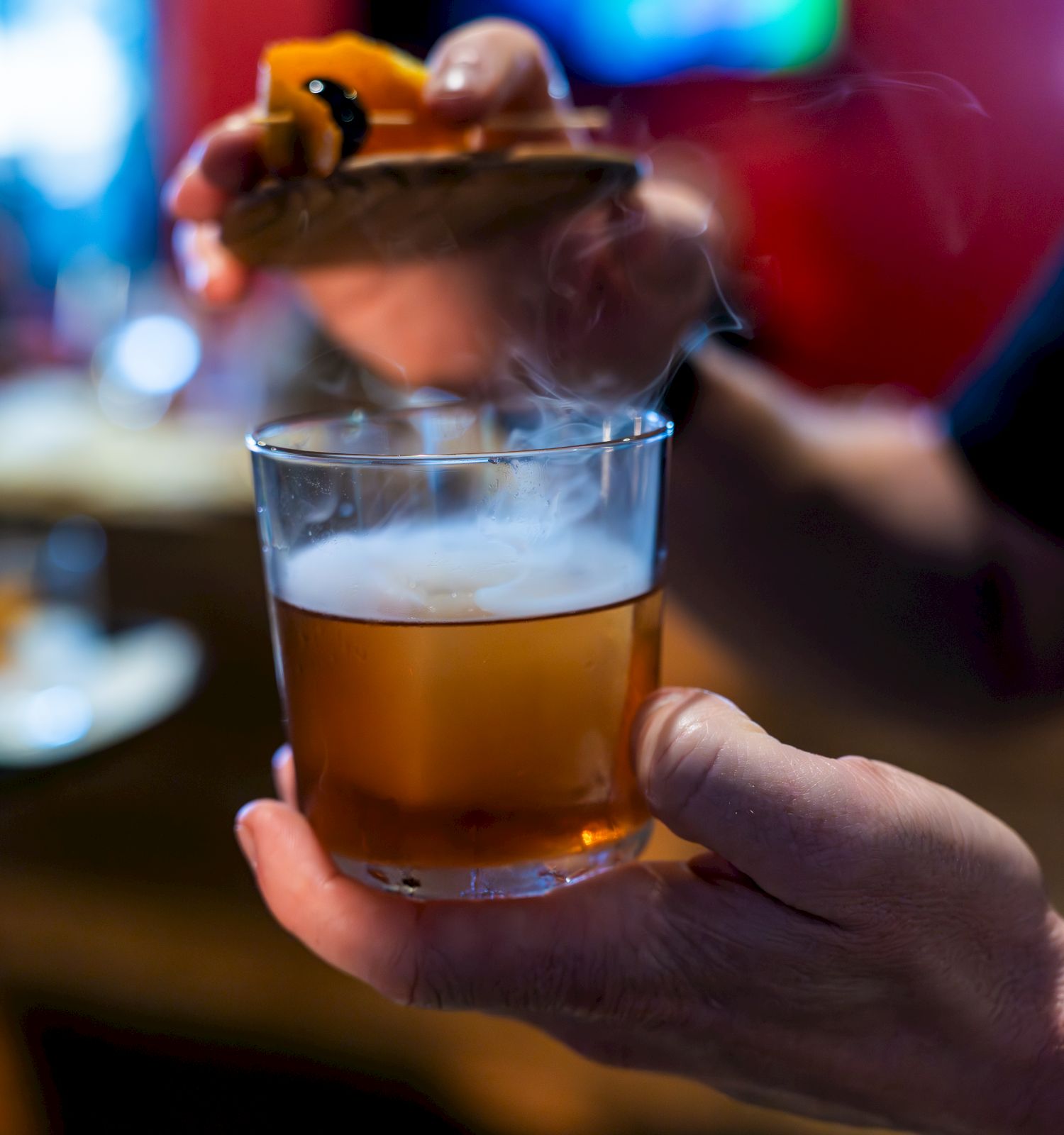 A hand holds a cocktail glass filled with a smoky beverage, while another hand places a smoky garnish on top.
