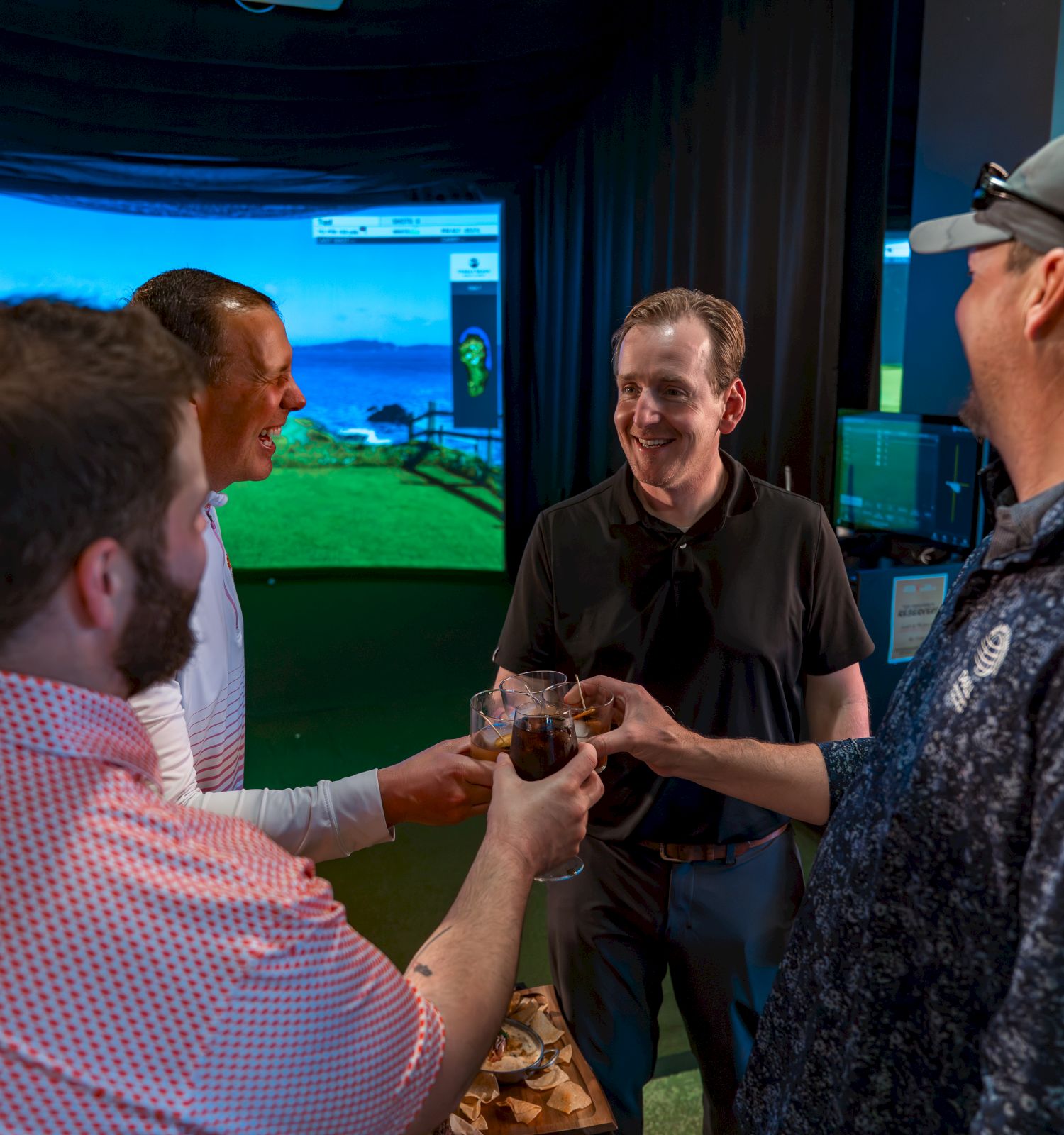 Four people are toasting drinks in an indoor setting with a virtual golf screen in the background.