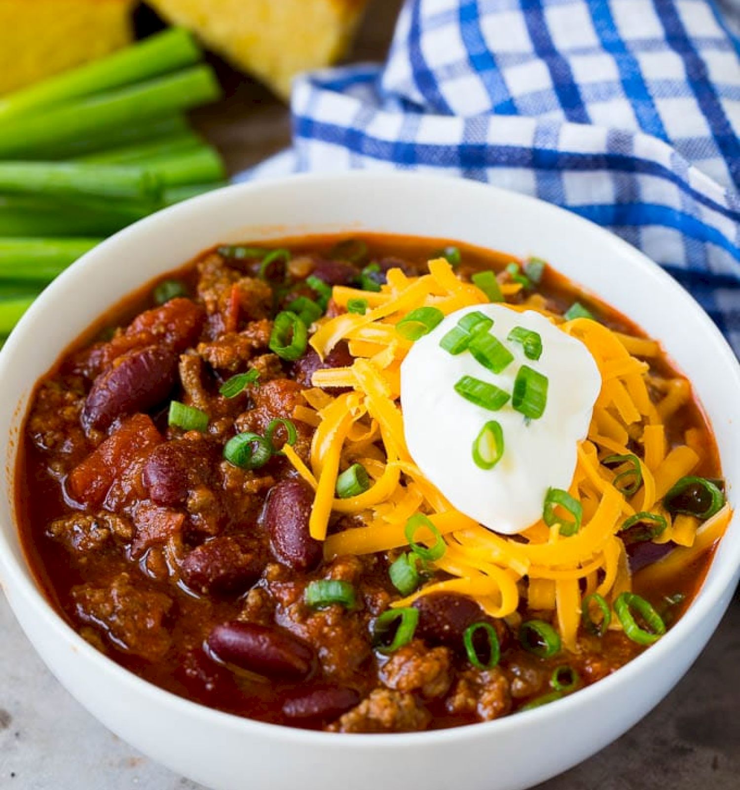 A bowl of chili topped with shredded cheese, sour cream, and green onions, with cornbread and a checkered cloth nearby.