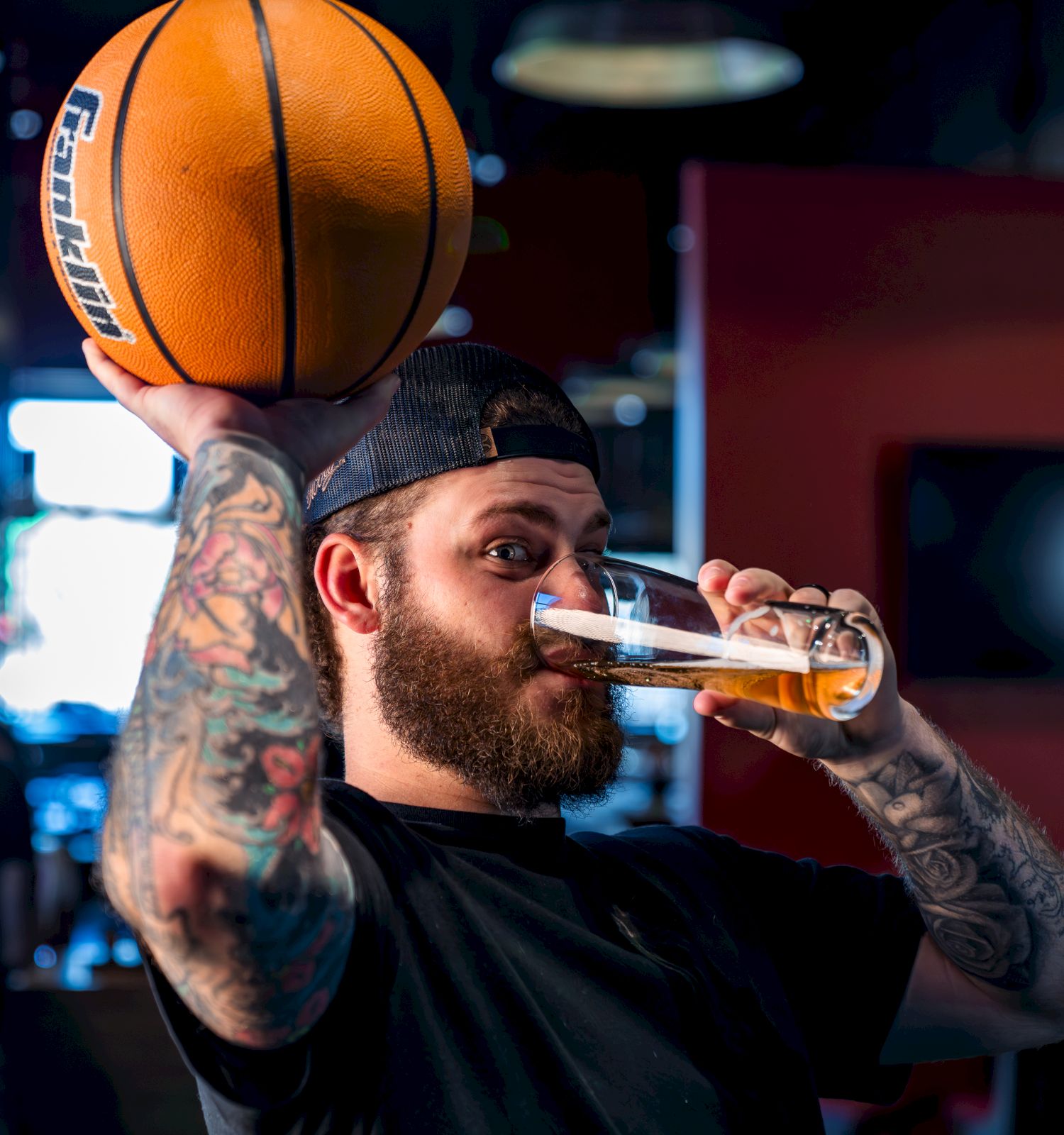 A man with tattoos holds a basketball and drinks from a glass in a dimly lit room.