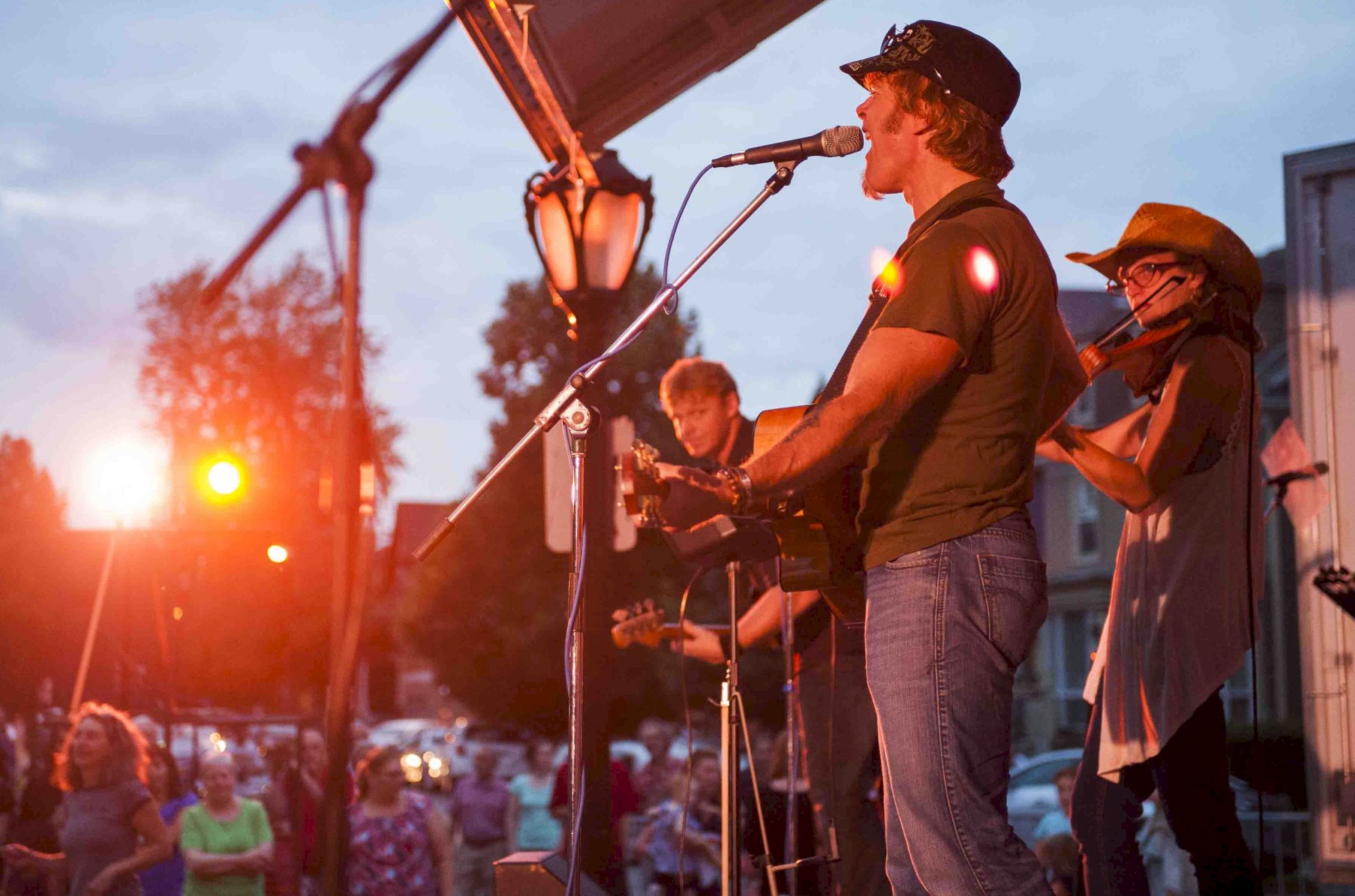 A band performs on an outdoor stage with a lively crowd and warm lighting, creating a vibrant, energetic atmosphere at dusk.