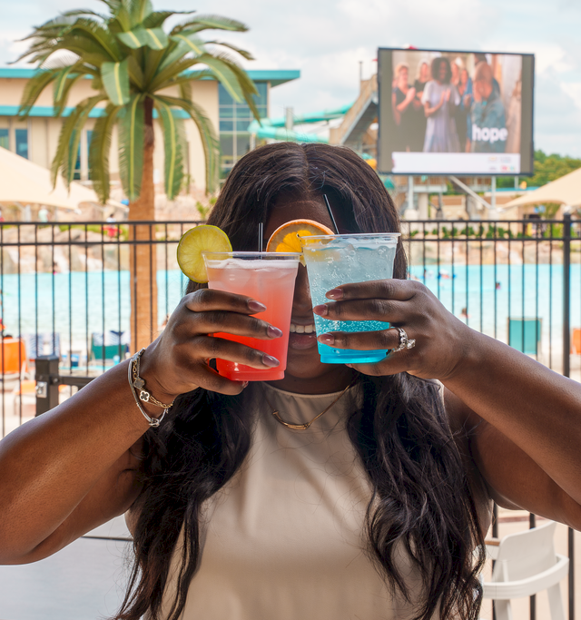 A woman by a pool bar clinks colorful cocktails with friends on a sunny day.