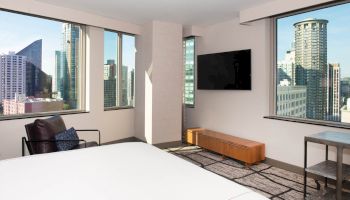 The image shows a modern hotel room with city views, large windows, a TV, bed, chair, and small table with an urban skyline backdrop.