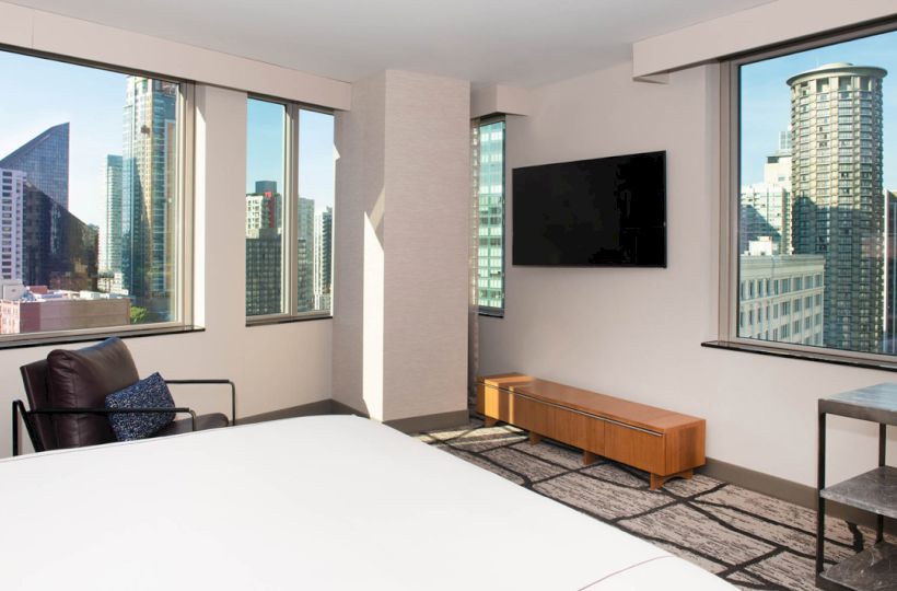 The image shows a modern hotel room with city views, large windows, a TV, bed, chair, and small table with an urban skyline backdrop.
