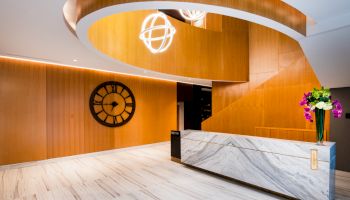 A modern lobby with a marble reception desk, large wall clock, stylish wood paneling, and hanging light fixtures.