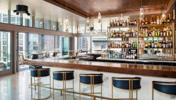 A modern bar with a sleek design, featuring a well-stocked liquor shelf and stylish stools, offering city views outside.