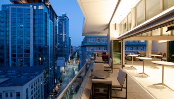 A modern rooftop terrace overlooks a cityscape with tall buildings, featuring stylish outdoor seating and a glass railing.