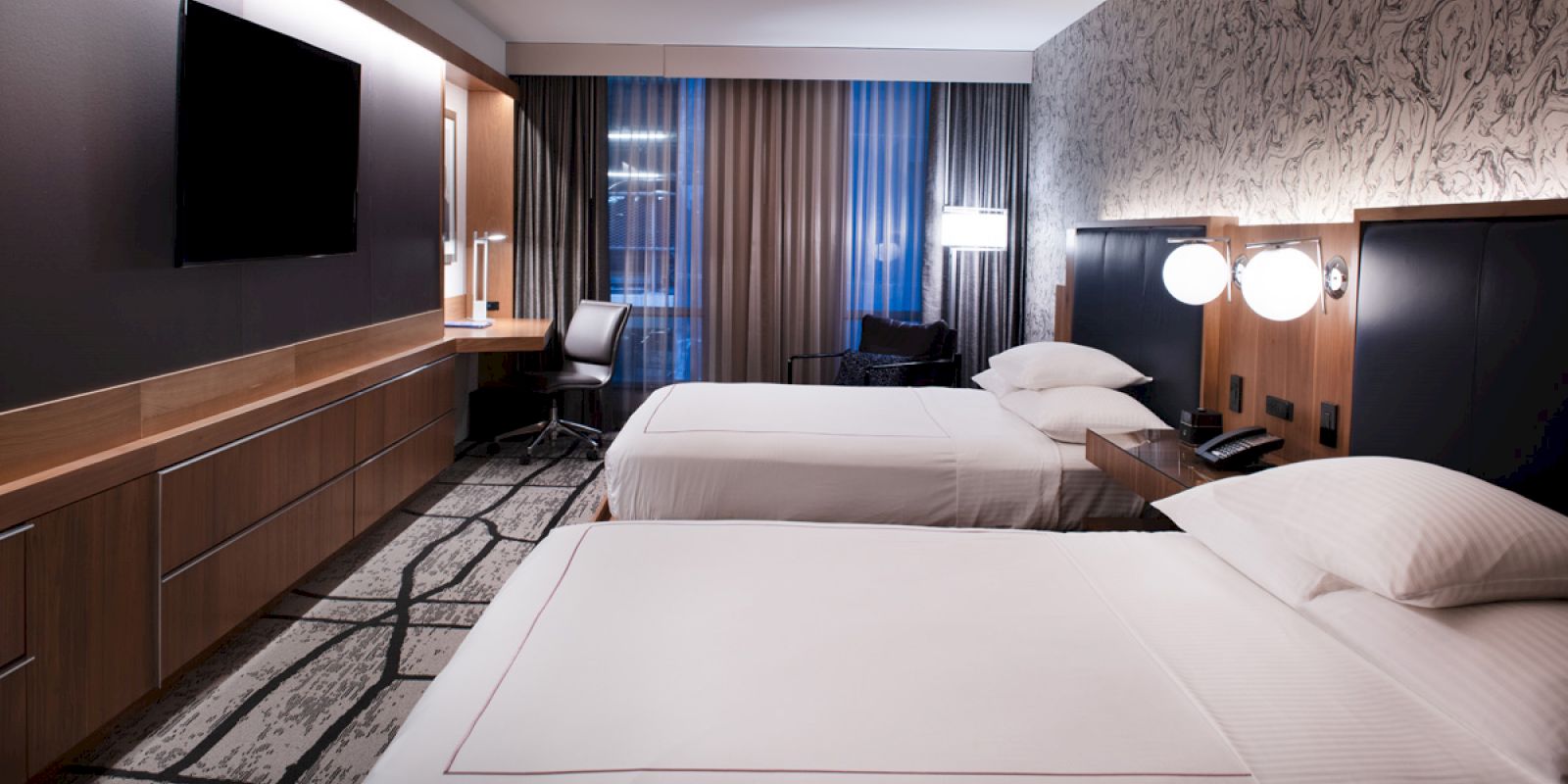 The image shows a modern hotel room with two beds, a TV, a desk, and contemporary lighting and decor, with large curtains covering windows.