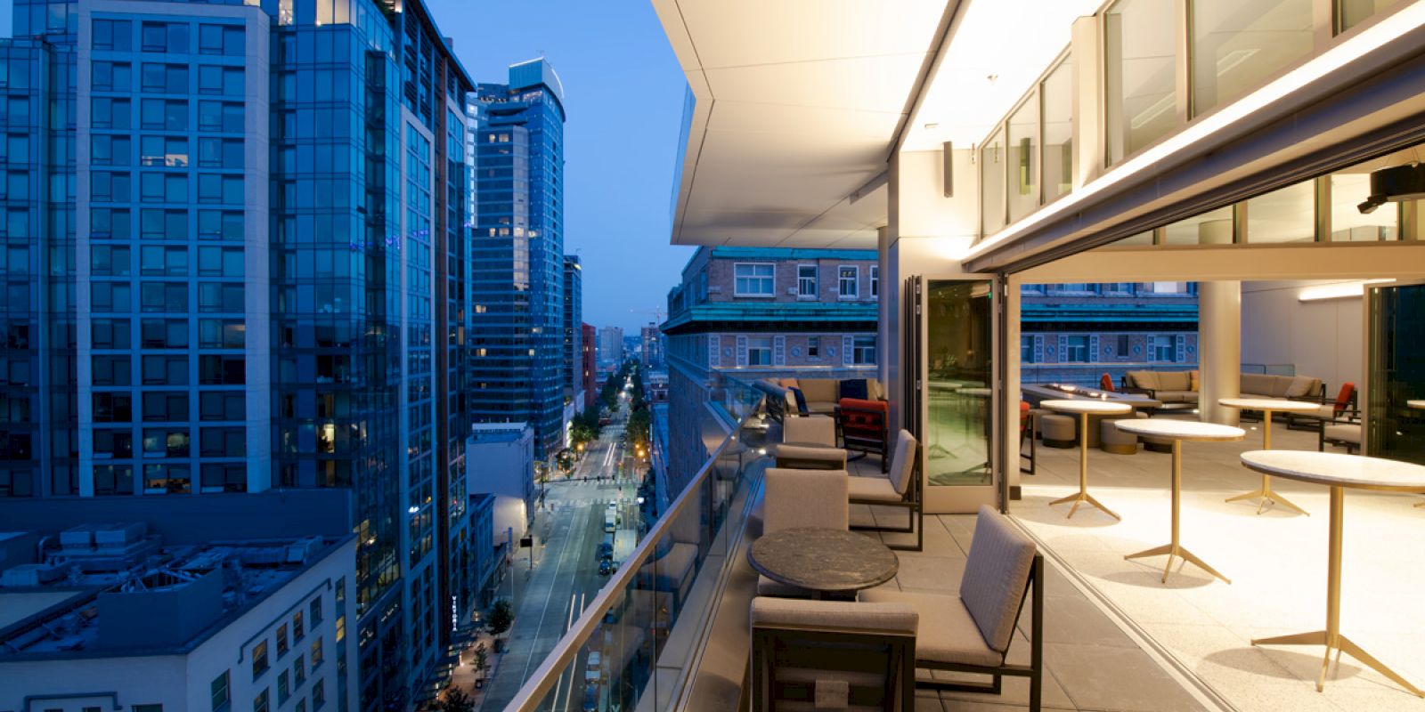 A rooftop terrace with seating overlooks a city street, surrounded by modern buildings under a twilight sky.