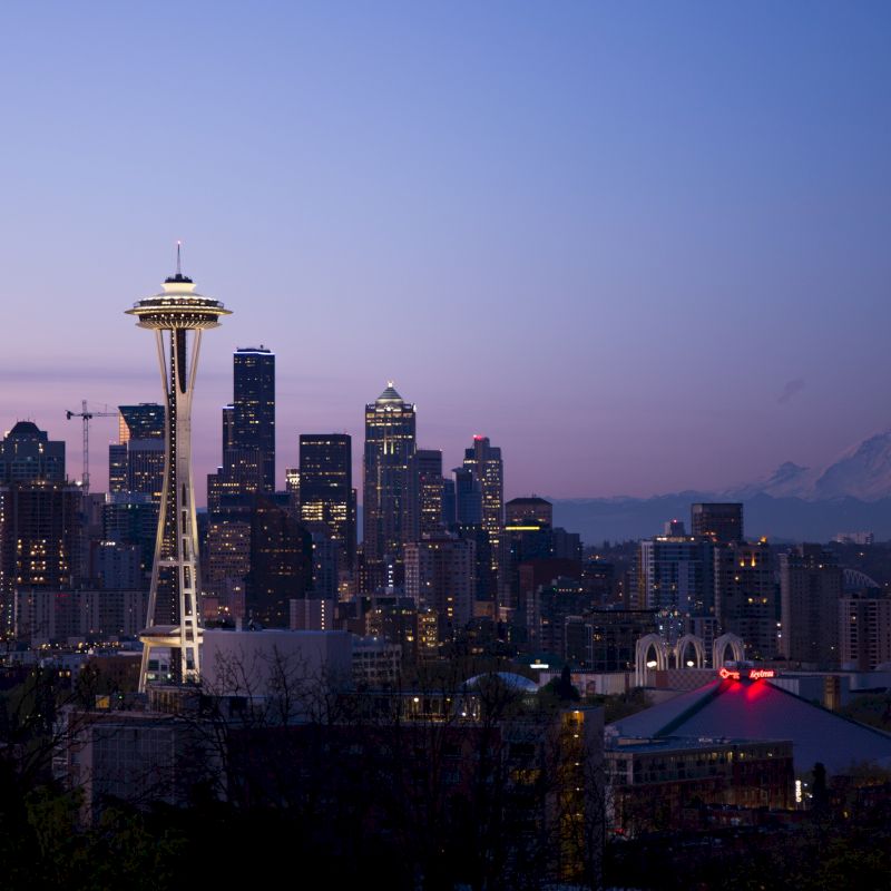 Seattle skyline at dusk with the Space Needle, city buildings, and Mount Rainier in the background against a purple sky.