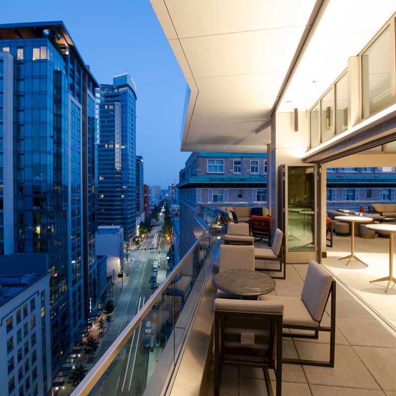 A modern rooftop terrace with seating overlooks a city street lined with tall buildings during the evening.
