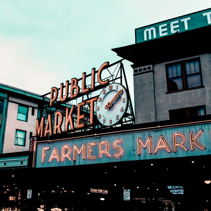 A neon-lit "Public Market" and "Farmers Market" sign with a clock in an urban setting, featuring vintage styled buildings.