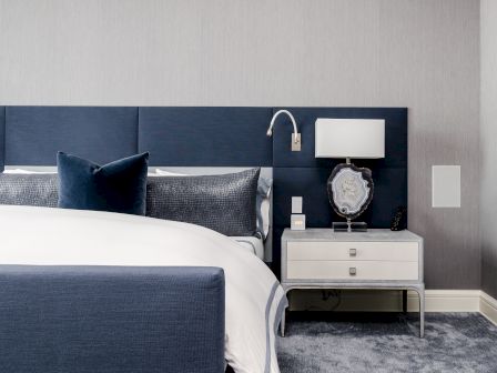 A modern bedroom with a navy blue bed, gray pillow, wall lamp, and a nightstand holding a decorative stone and white lamp.
