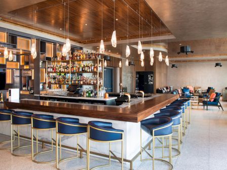 A modern bar with blue stools, a well-stocked back shelf, pendant lights, and lounge seating in the background in a stylish setting.
