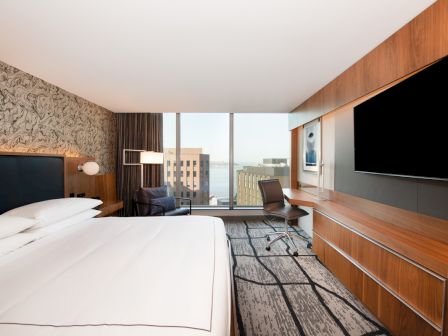 A modern hotel room with a large bed, wall-mounted TV, desk, chair, and a window view of buildings and water in the background.
