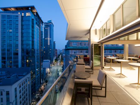 A modern balcony with seating overlooks an urban street lined with tall buildings under a twilight sky.