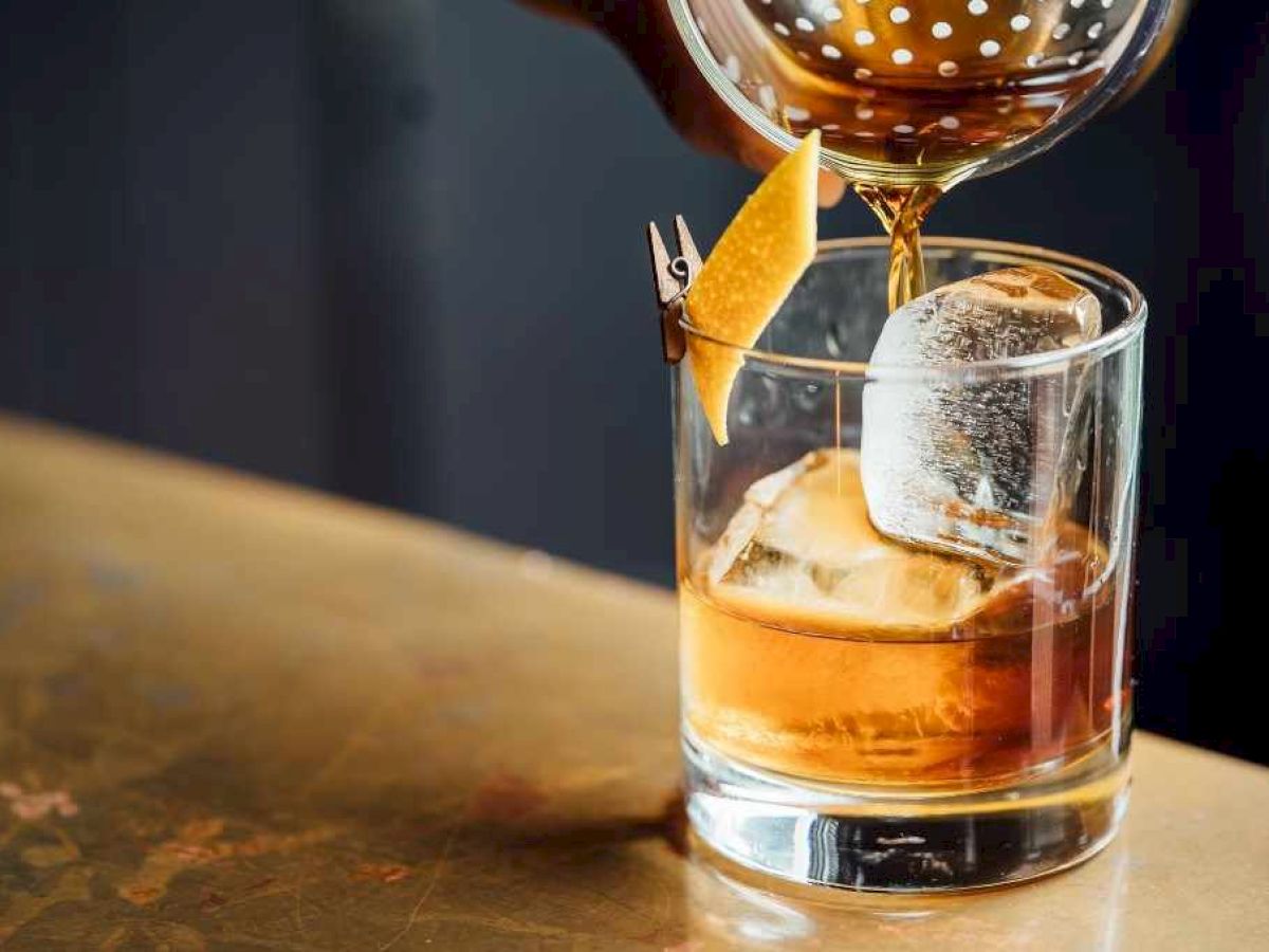 A cocktail is being poured into a glass with ice and a citrus twist on the rim.