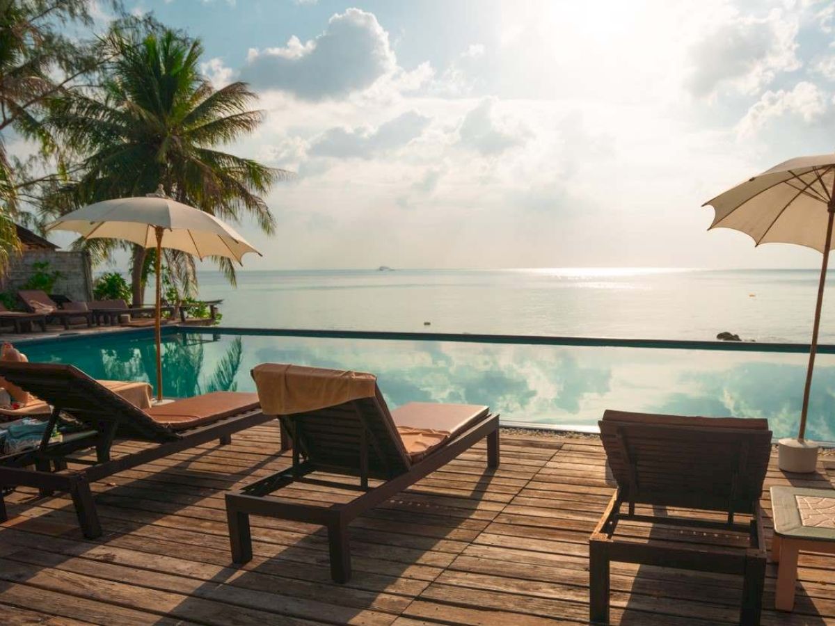 A serene poolside setting with lounge chairs, umbrellas, palm trees, and a view of the ocean under a partly cloudy sky.