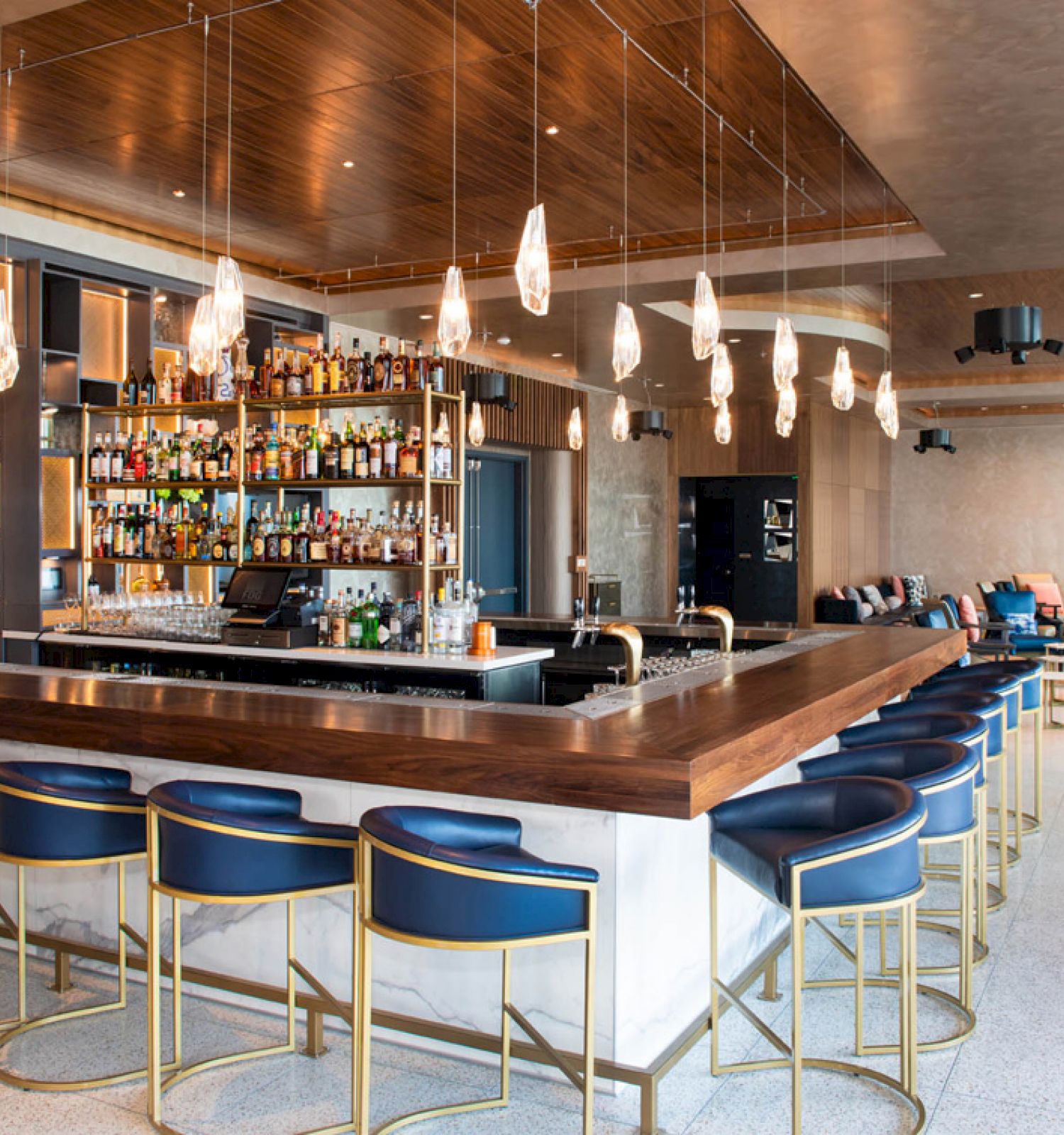 The image shows a modern bar with stylish seating, a well-stocked liquor display, hanging lights, and a cozy lounge area in the background.