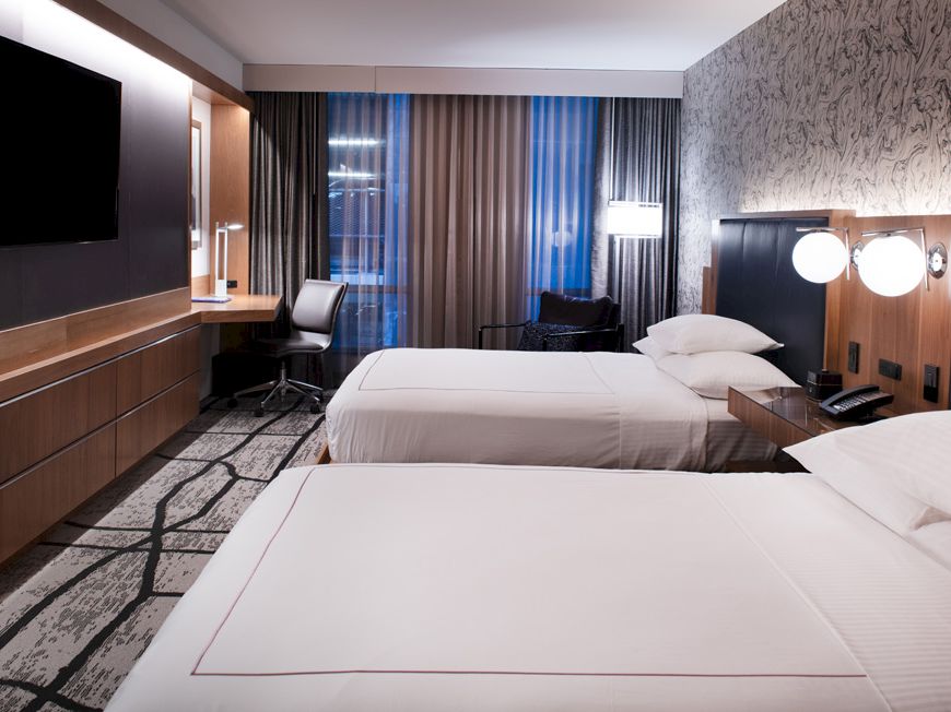A modern hotel room with two beds, a wall-mounted TV, desk, chair, and curtains. The room has a contemporary design and lighting.