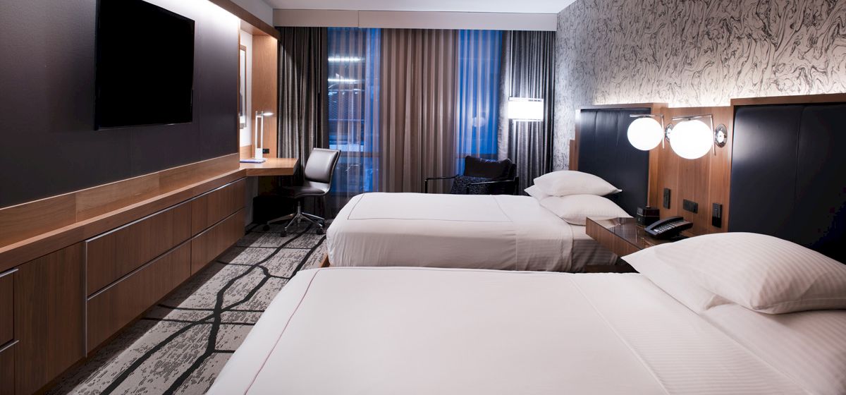 A modern hotel room with two beds, a wall-mounted TV, desk, chair, and curtains. The room has a contemporary design and lighting.