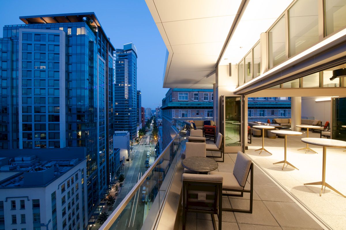 A modern outdoor terrace with seating overlooks a city street lined with tall glass buildings at dusk.