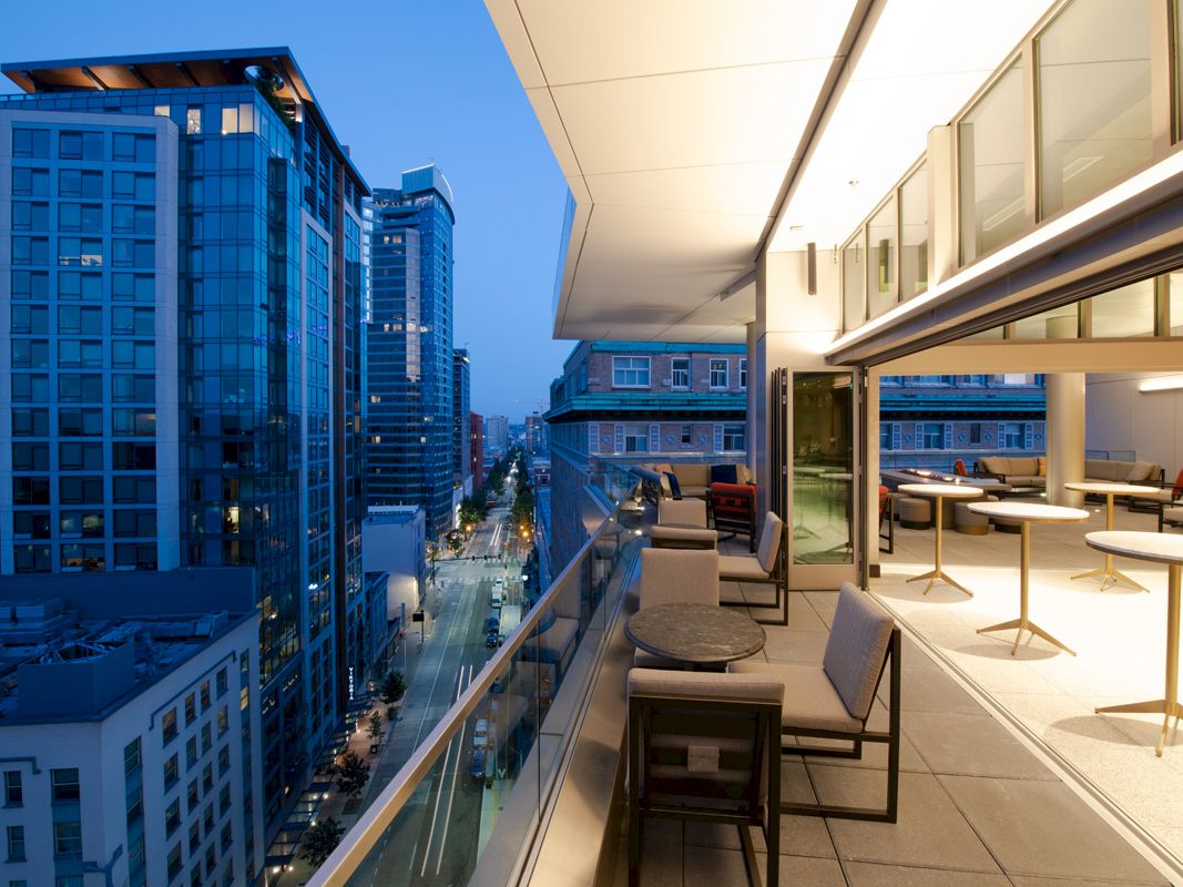 A modern outdoor terrace with seating overlooks a city street lined with tall glass buildings at dusk.
