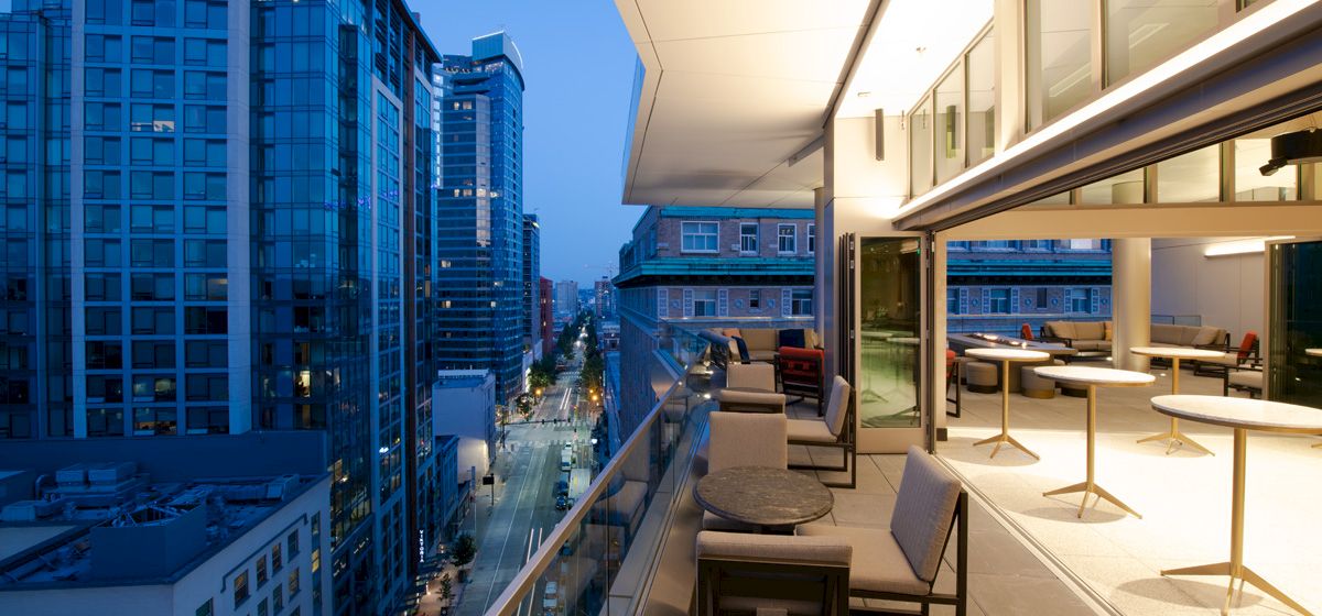A modern outdoor terrace with seating overlooks a city street lined with tall glass buildings at dusk.