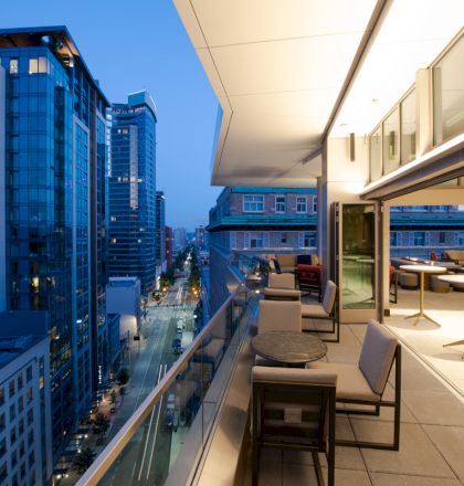 A modern cityscape at dusk with a view from an upscale balcony featuring seating, overlooking lit skyscrapers and streets below.