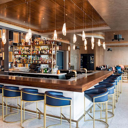 A modern bar with blue stools, a stocked liquor display, pendant lights, and a spacious seating area.