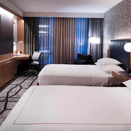A modern hotel room with two beds, a TV on the wall, a desk with a chair, and lamps on the nightstand.