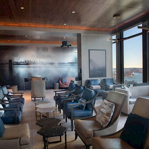 A modern lounge with stylish seating, floor-to-ceiling windows offering a scenic view, and a wall mural of a skyline in the background.