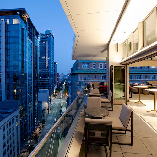 A cityscape view from a modern balcony at twilight, featuring tall buildings, outdoor seating, and bright indoor lighting.