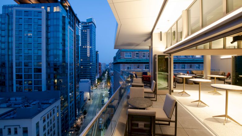 A modern rooftop terrace with seating overlooks a city street flanked by tall buildings, lit by evening lights.