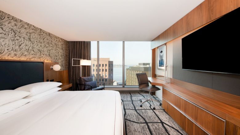 A modern hotel room with a large bed, desk, chair, and TV. There's a view outside the window with buildings and some water visible.