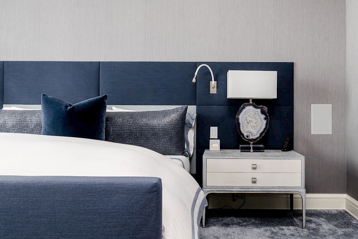 The image shows a modern bedroom with a navy and white color scheme, featuring a bed, cushions, side table, lamp, and wall light fixture.