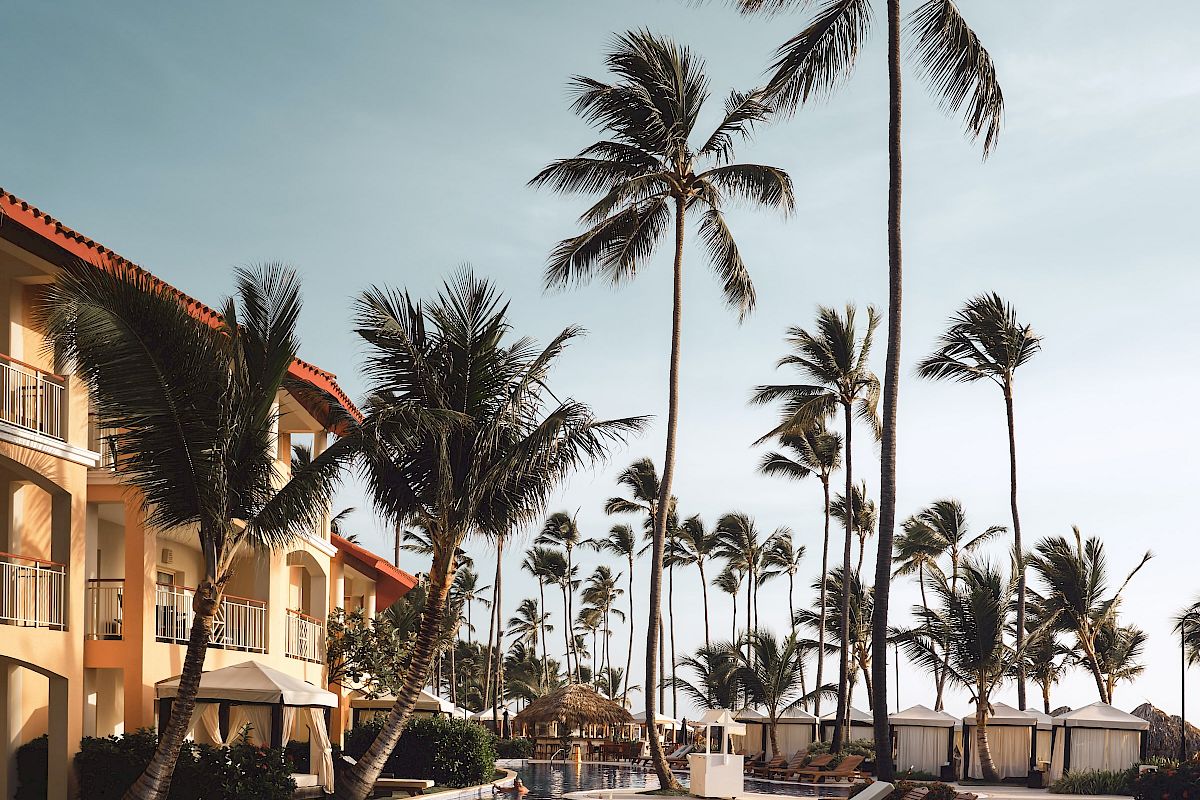 A tropical resort scene with a pool, lounge chairs, and tall palm trees under a clear sky, next to a beige building with balconies.