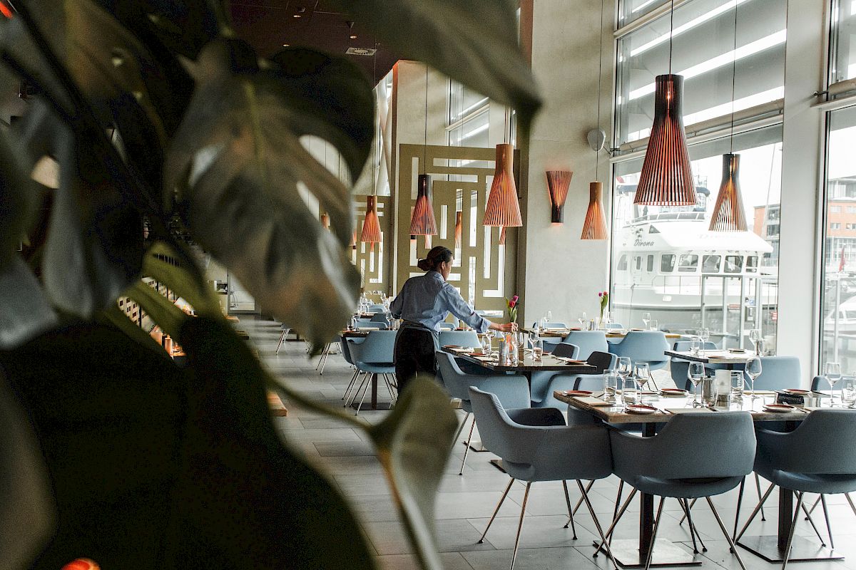 A person setting tables in a modern restaurant with large windows and stylish decor, featuring hanging lights and plant leaves in view.