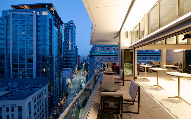 A modern balcony with seating overlooks a cityscape featuring tall buildings, with the scene captured at dusk.