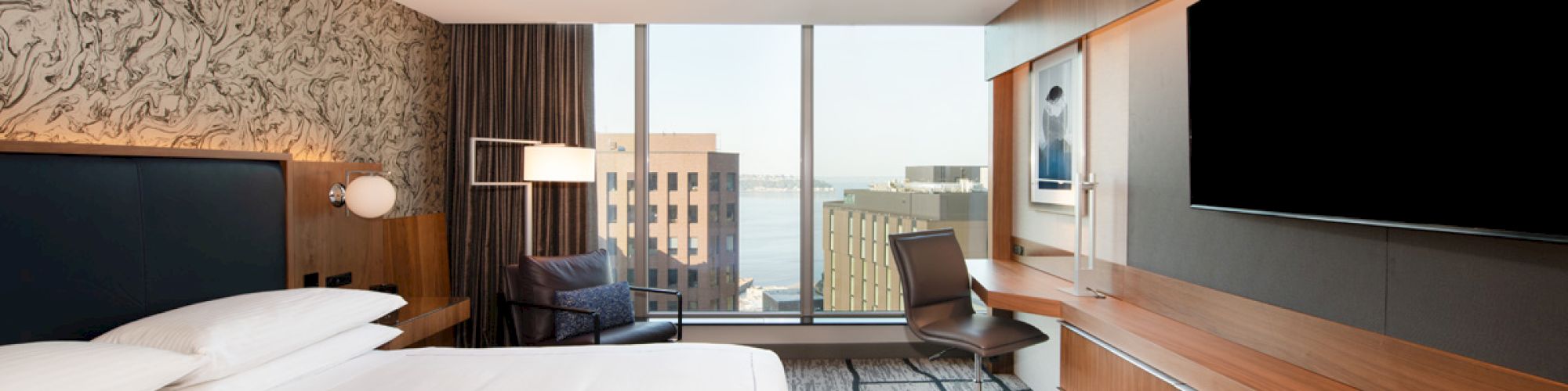 The image shows a modern hotel room with a large bed, desk, chair, TV, and a window offering a city view.